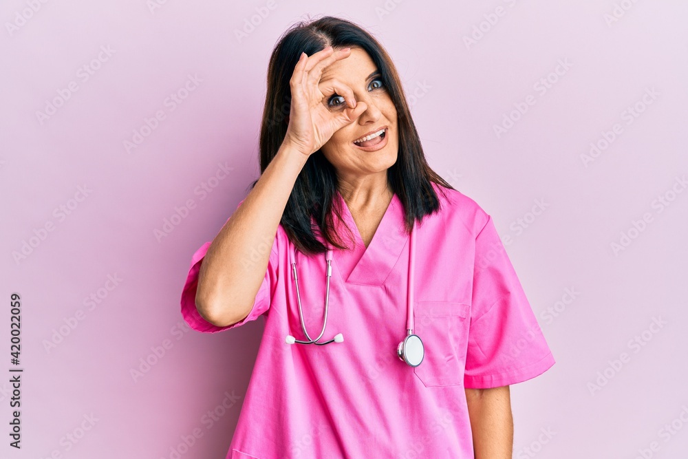 Middle age brunette woman wearing doctor uniform and stethoscope doing ok gesture with hand smiling, eye looking through fingers with happy face. Middle age brunette woman wearing doctor uniform and stethoscope doing ok gesture with hand smiling, eye looking through fingers with happy face.