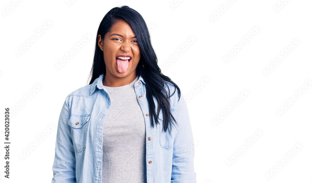 Hispanic woman with long hair wearing casual denim jacket sticking tongue out happy with funny expression. emotion concept. Hispanic woman with long hair wearing casual denim jacket sticking tongue out happy with funny expression. emotion concept.
