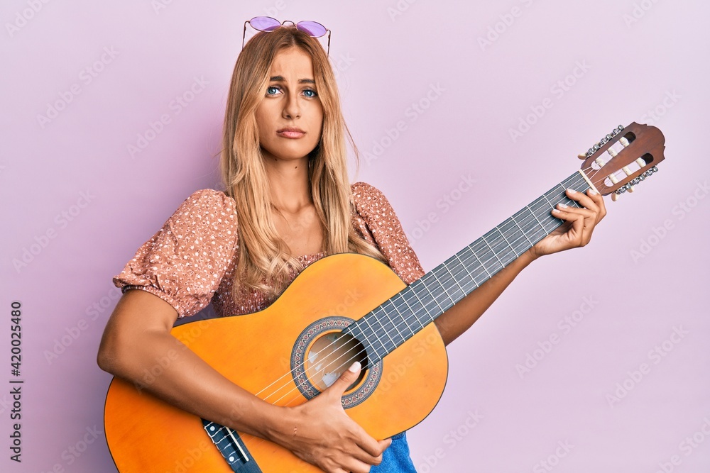 Fototapeta premium Beautiful blonde young woman playing classical guitar clueless and confused expression. doubt concept.