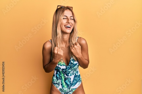 Beautiful blonde young woman wearing swimsuit and sunglasses very happy and excited doing winner gesture with arms raised, smiling and screaming for success. celebration concept.