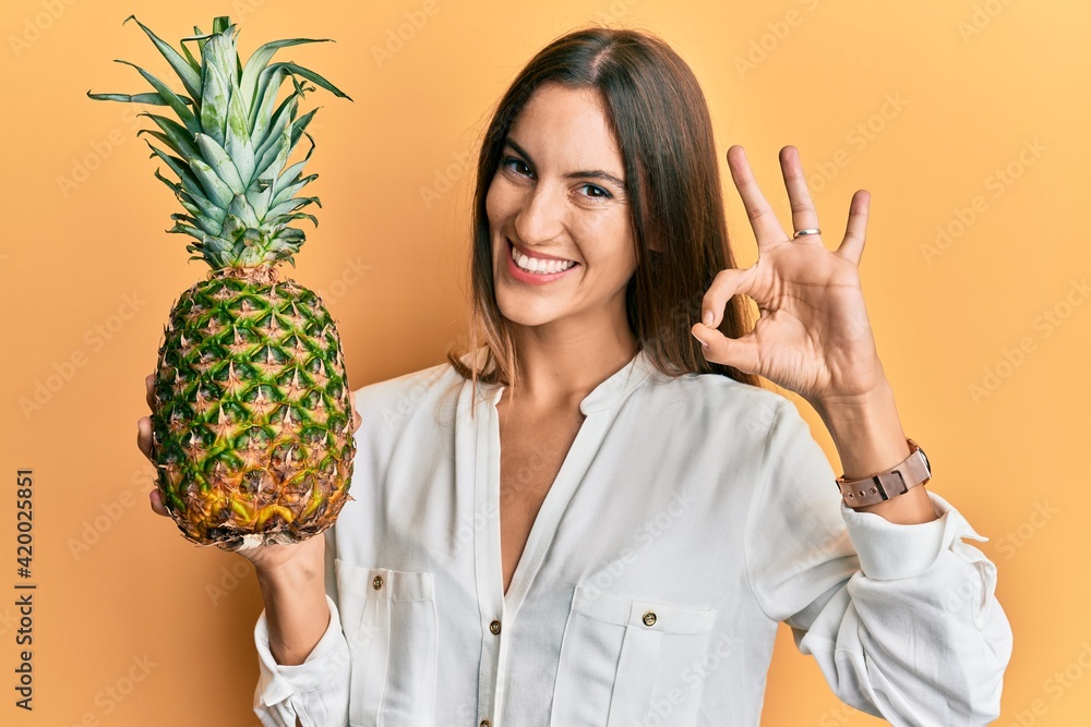 Young beautiful woman holding pineapple doing ok sign with fingers, smiling friendly gesturing excellent symbol Young beautiful woman holding pineapple doing ok sign with fingers, smiling friendly gesturing excellent symbol
