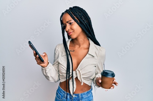 Beautiful hispanic woman using smartphone and drinking a cup of coffee skeptic and nervous, frowning upset because of problem. negative person.