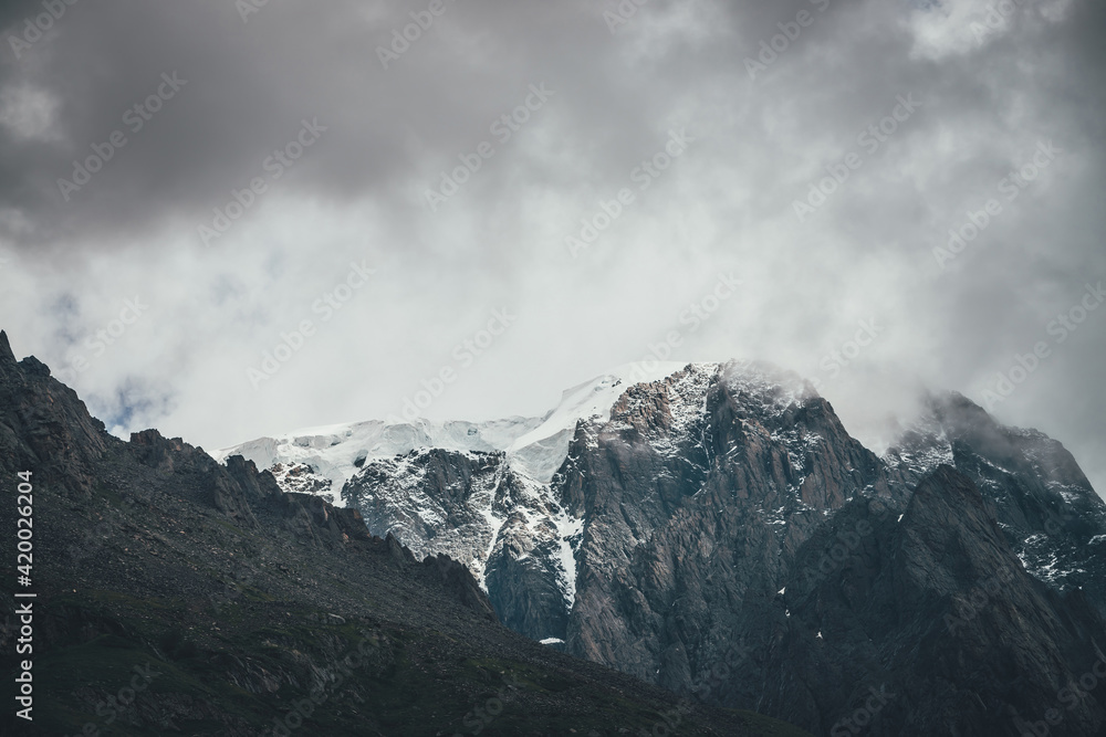 Dark atmospheric surreal landscape with dark rocky mountain top in low clouds in gray cloudy sky. Gray low cloud on high pinnacle. High black rock with snow in low clouds. Surrealist gloomy mountains.