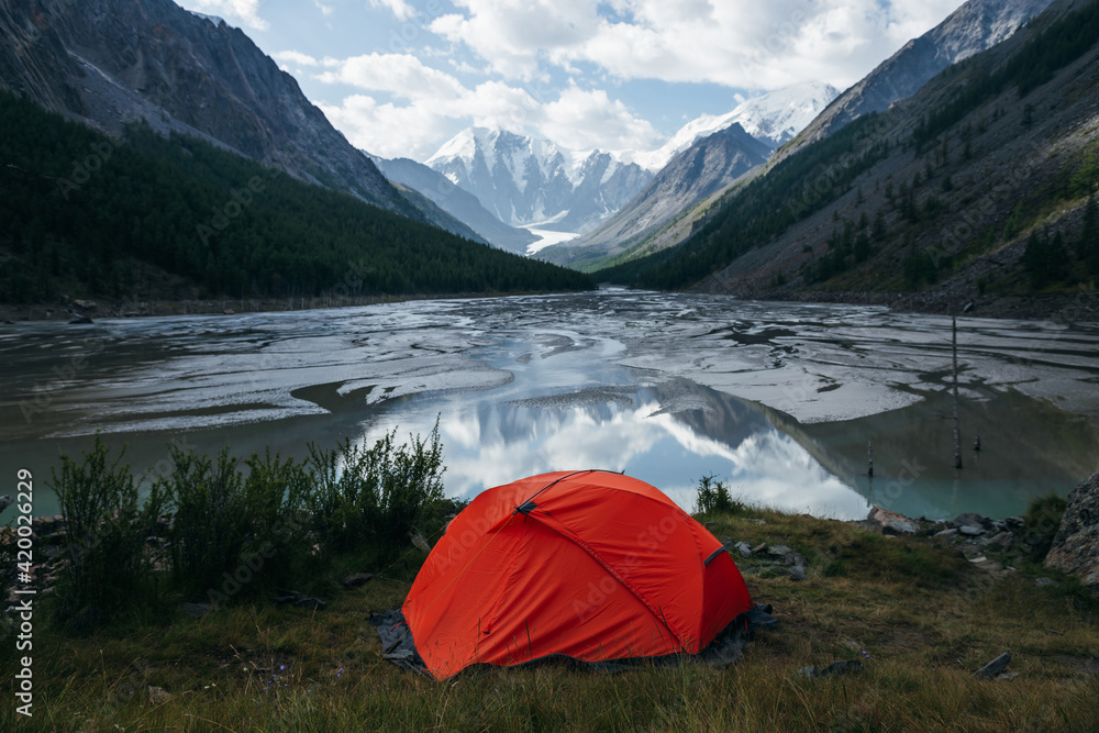 Scenic alpine landscape with orange tent near beautiful mirror mountain lake with streams in highland valley from snowy mountains under cloudy sky. Sunny high mountain scenery with mirror glacial lake