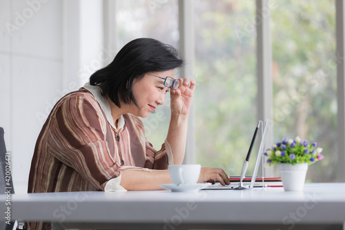 Asian older woman wearing eyeglasses working with a laptop computer with presbyopia longsighted eyes problem and fell stessed photo