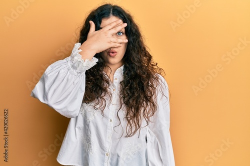 Young brunette woman with curly hair wearing casual clothes peeking in shock covering face and eyes with hand, looking through fingers afraid