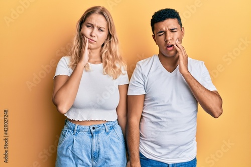 Young interracial couple wearing casual white tshirt touching mouth with hand with painful expression because of toothache or dental illness on teeth. dentist