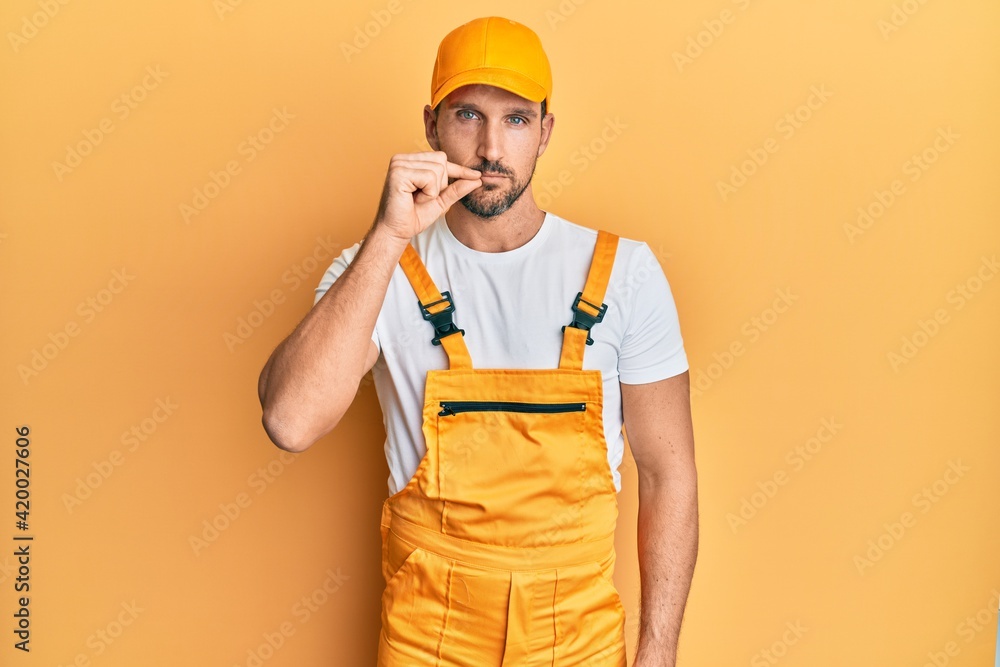 Young handsome man wearing handyman uniform over yellow background mouth and lips shut as zip with fingers. secret and silent, taboo talking Young handsome man wearing handyman uniform over yellow background mouth and lips shut as zip with fingers. secret and silent, taboo talking