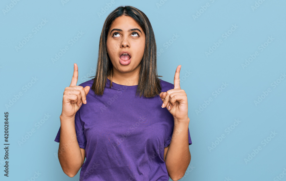 Young hispanic girl wearing casual purple t shirt amazed and surprised looking up and pointing with fingers and raised arms. Young hispanic girl wearing casual purple t shirt amazed and surprised looking up and pointing with fingers and raised arms.
