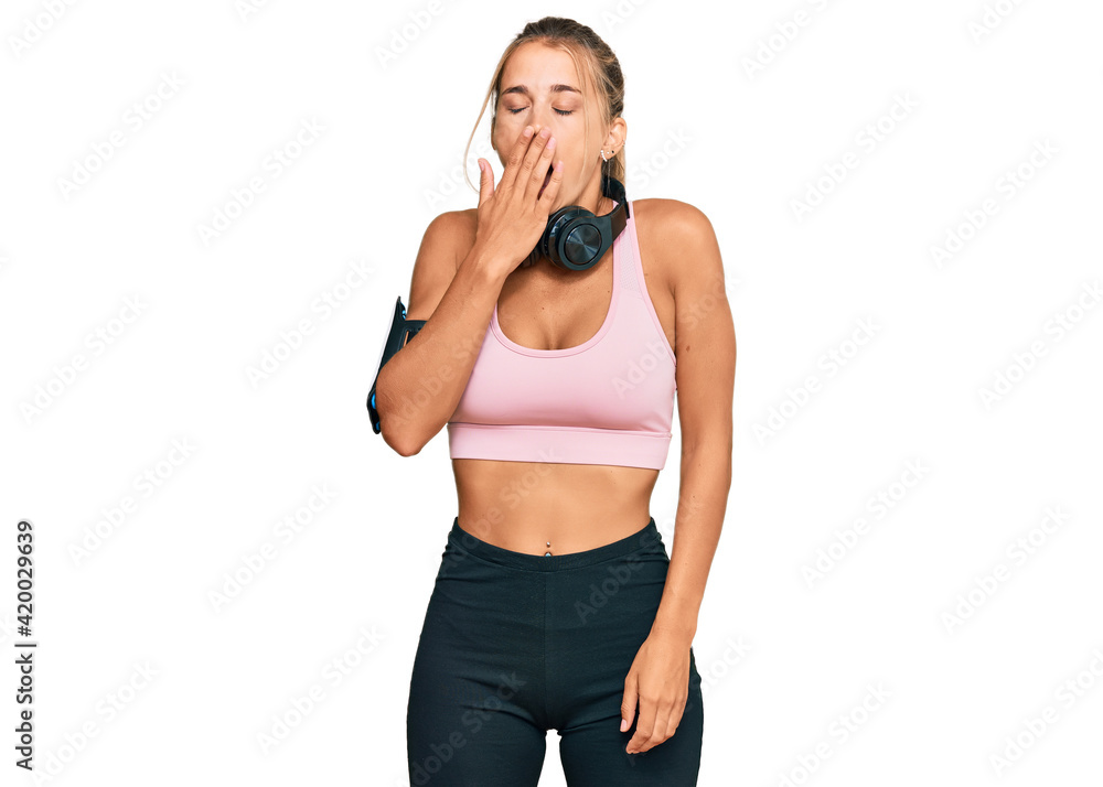 Young blonde woman wearing gym clothes and using headphones bored yawning tired covering mouth with hand. restless and sleepiness. Young blonde woman wearing gym clothes and using headphones bored yawning tired covering mouth with hand. restless and sleepiness.