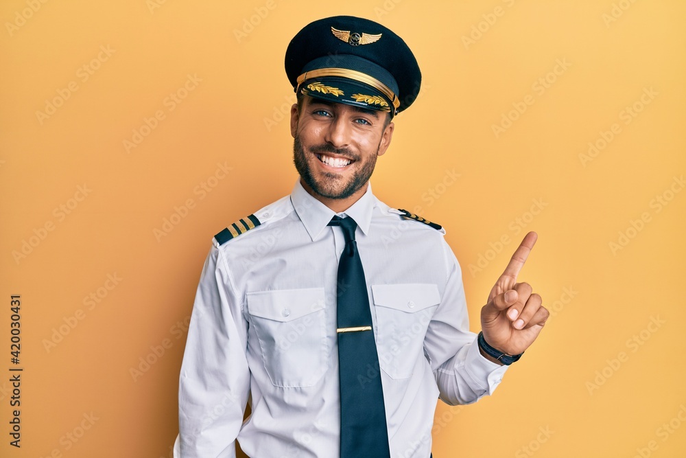 Handsome hispanic man wearing airplane pilot uniform with a big smile on face, pointing with hand finger to the side looking at the camera. - obrazy, fototapety, plakaty 