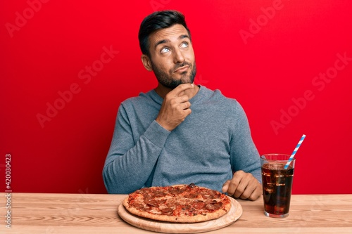 Handsome hispanic man eating tasty pepperoni pizza thinking concentrated about doubt with finger on chin and looking up wondering