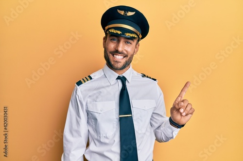 Fotobehang Handsome hispanic man wearing airplane pilot uniform with a big smile on face, pointing with hand finger to the side looking at the camera