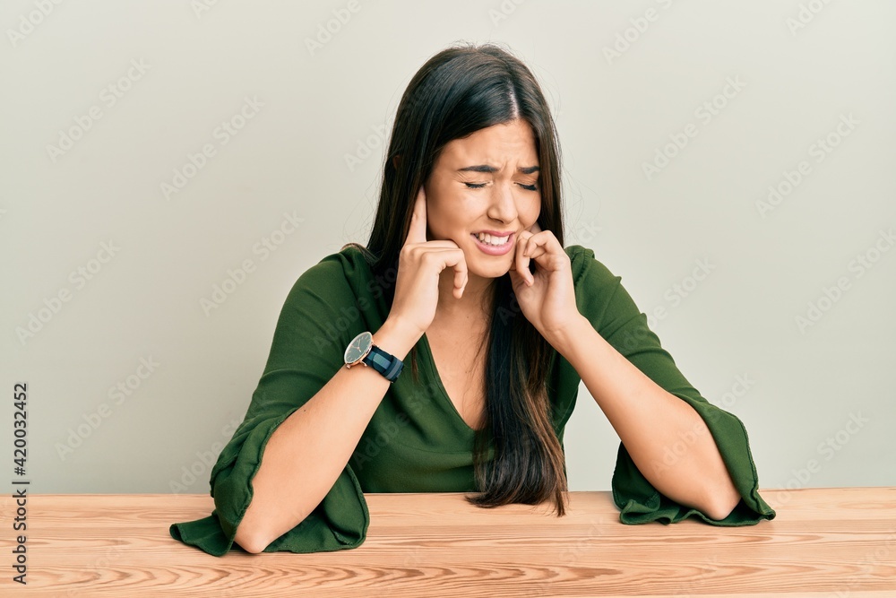 Young brunette woman wearing casual clothes sitting on the table covering ears with fingers with annoyed expression for the noise of loud music. deaf concept. Young brunette woman wearing casual clothes sitting on the table covering ears with fingers with annoyed expression for the noise of loud music. deaf concept.