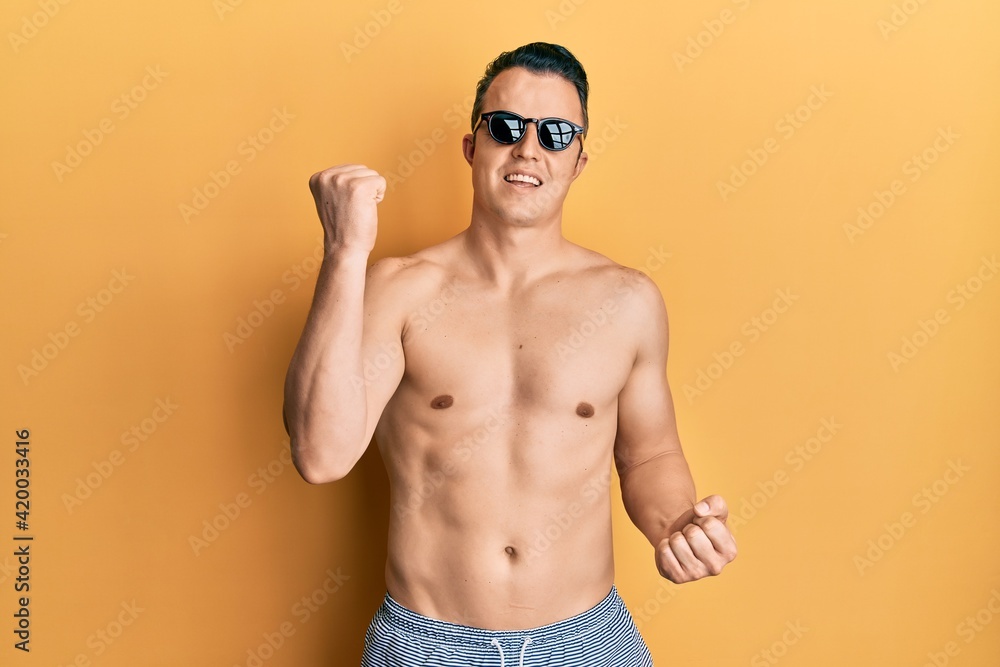 Handsome young man wearing swimsuit and sunglasses celebrating surprised and amazed for success with arms raised and eyes closed