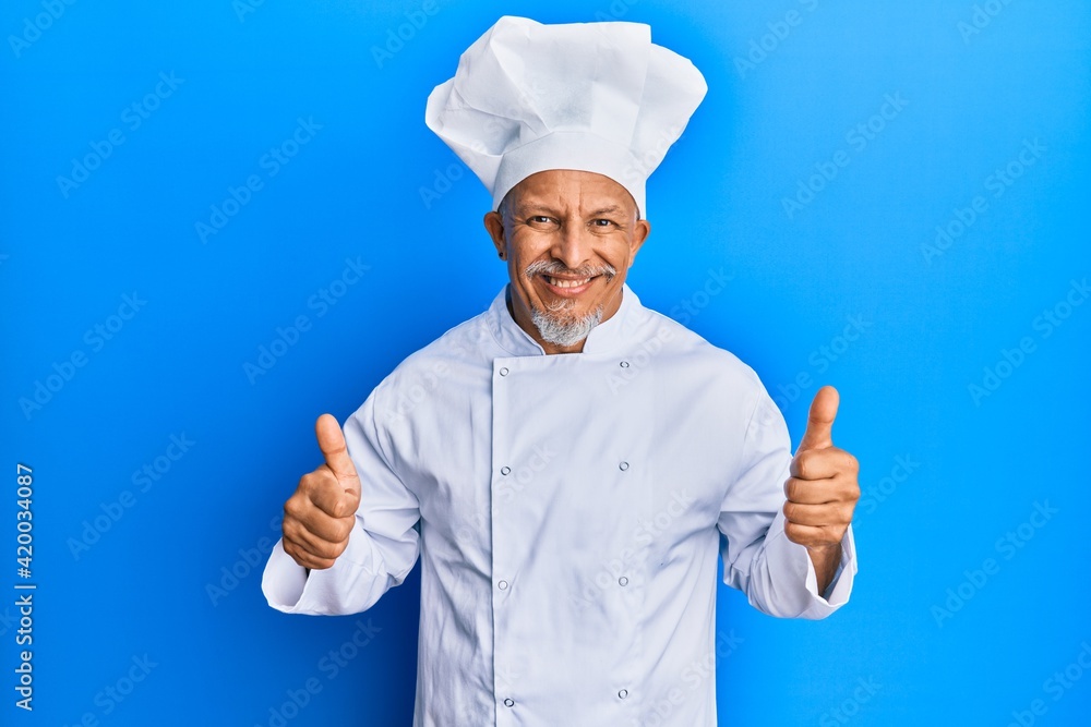 Middle age grey-haired man wearing professional cook uniform and hat success sign doing positive gesture with hand, thumbs up smiling and happy. cheerful expression and winner gesture. Middle age grey-haired man wearing professional cook uniform and hat success sign doing positive gesture with hand, thumbs up smiling and happy. cheerful expression and winner gesture.