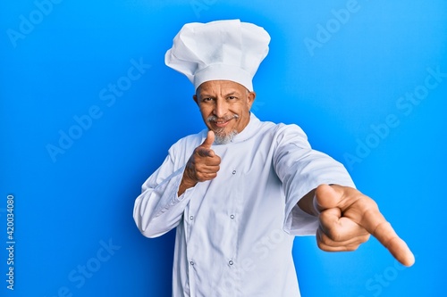 Middle age grey-haired man wearing professional cook uniform and hat pointing fingers to camera with happy and funny face. good energy and vibes.