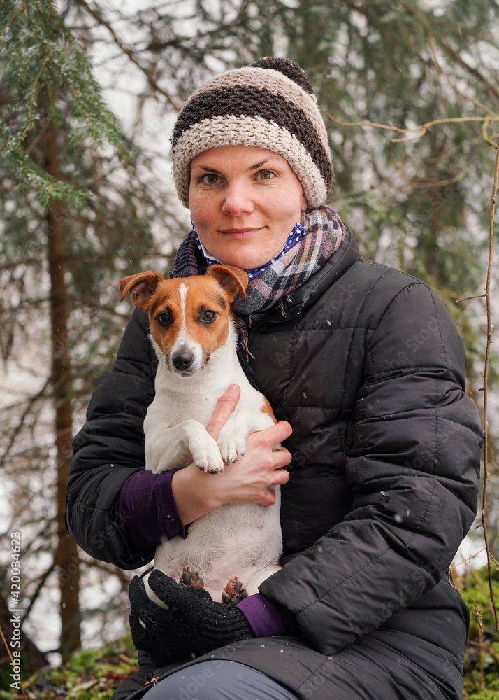 Young woman in winter jacket holding her Jack Russell terrier dog on hands, blurred trees background Young woman in winter jacket holding her Jack Russell terrier dog on hands, blurred trees background
