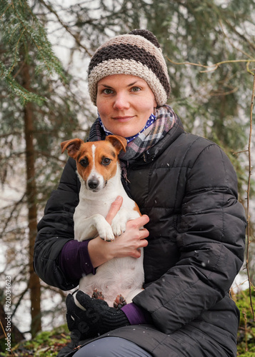 Young woman in winter jacket holding her Jack Russell terrier dog on hands, blurred trees background © Lubo Ivanko
