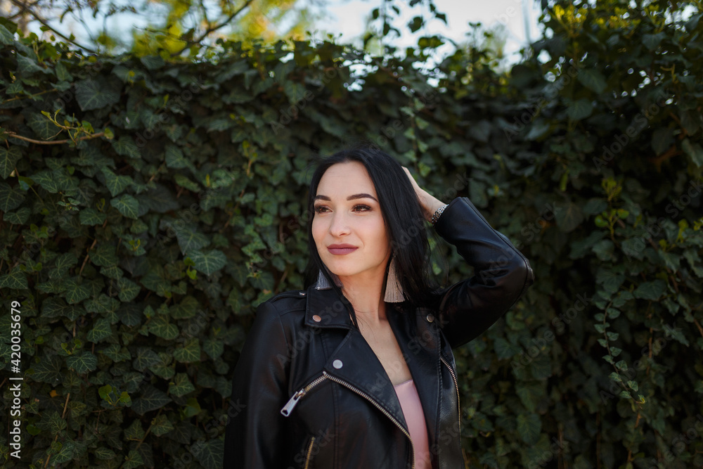 Portrait of young pretty dark-haired woman in black leather jacket posing on green ivy plant background. Portrait of young pretty dark-haired woman in black leather jacket posing on green ivy plant background.