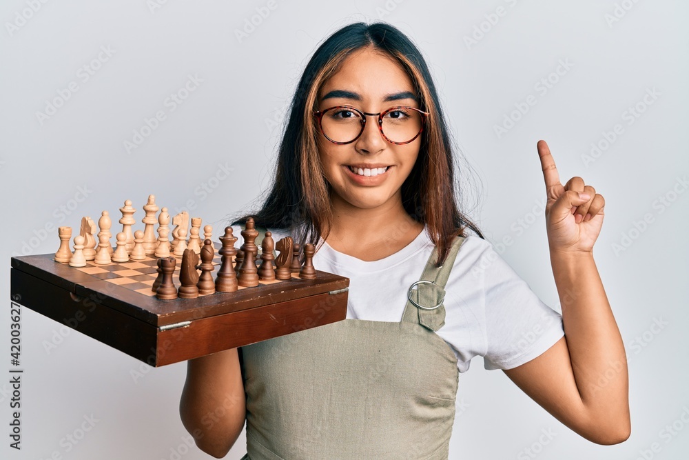 Young latin woman holding chess smiling with an idea or question pointing finger with happy face, number one Young latin woman holding chess smiling with an idea or question pointing finger with happy face, number one