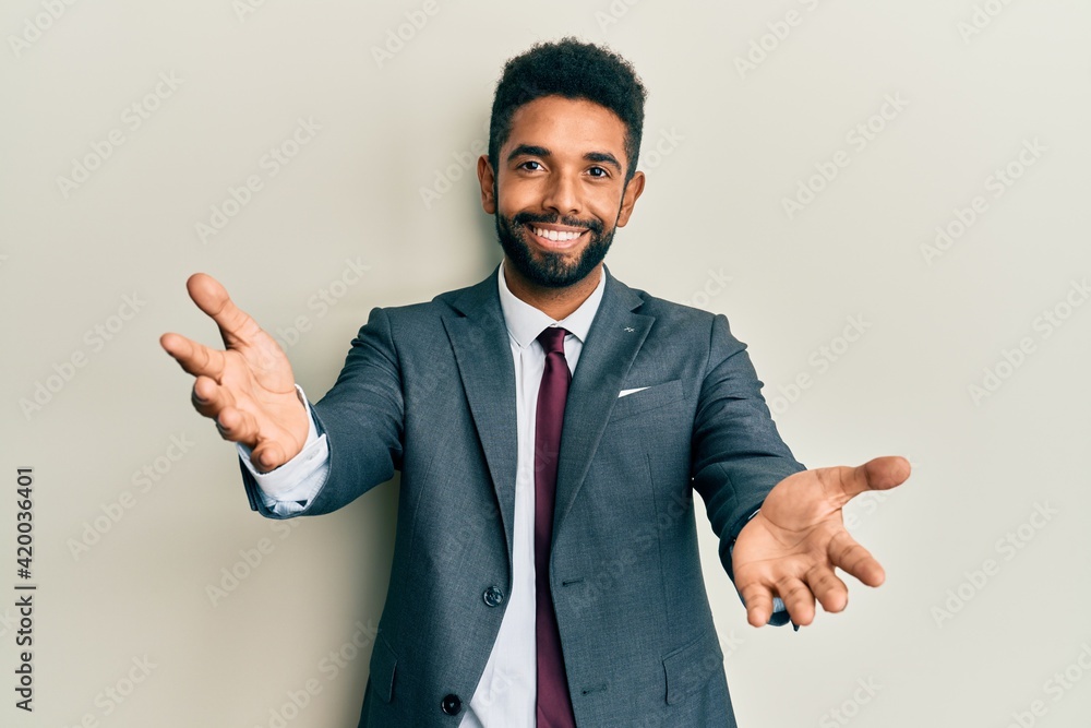 Handsome hispanic man with beard wearing business suit and tie looking at the camera smiling with open arms for hug. cheerful expression embracing happiness. Handsome hispanic man with beard wearing business suit and tie looking at the camera smiling with open arms for hug. cheerful expression embracing happiness.