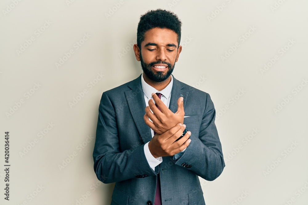Handsome hispanic man with beard wearing business suit and tie suffering pain on hands and fingers, arthritis inflammation Handsome hispanic man with beard wearing business suit and tie suffering pain on hands and fingers, arthritis inflammation
