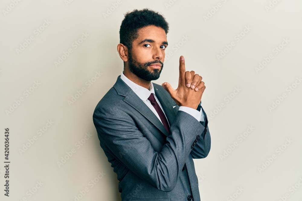 Handsome hispanic man with beard wearing business suit and tie holding symbolic gun with hand gesture, playing killing shooting weapons, angry face Handsome hispanic man with beard wearing business suit and tie holding symbolic gun with hand gesture, playing killing shooting weapons, angry face