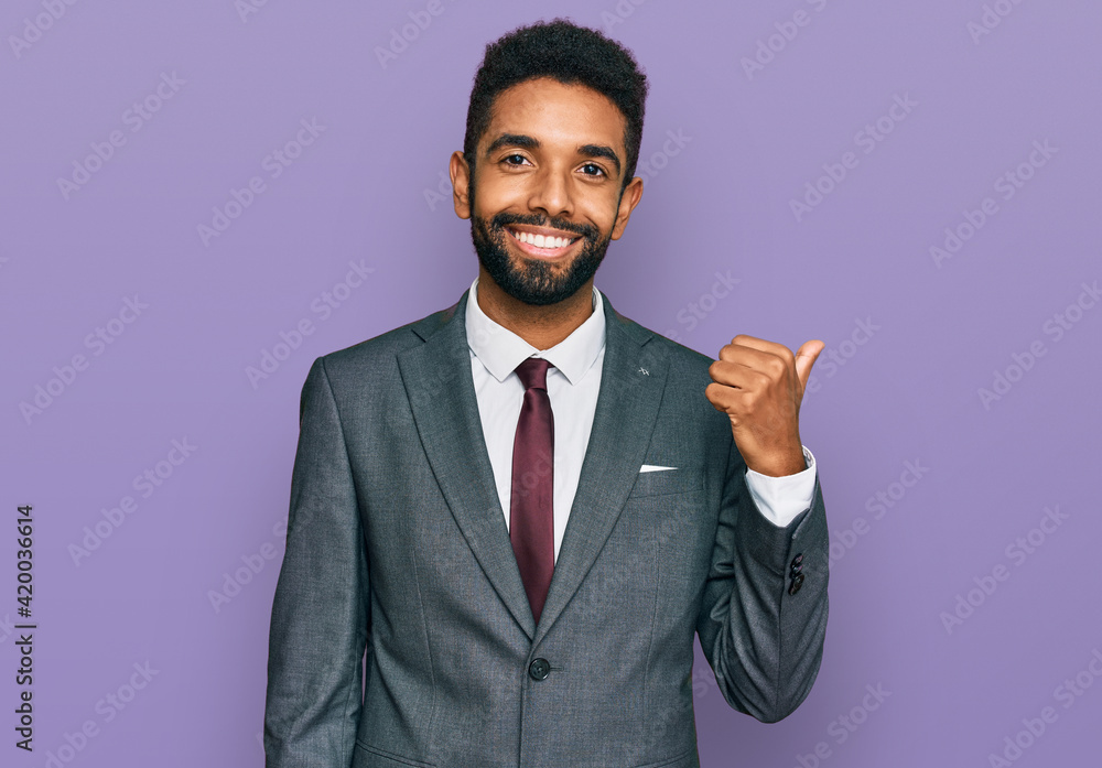 Young african american man wearing business clothes smiling with happy face looking and pointing to the side with thumb up. Young african american man wearing business clothes smiling with happy face looking and pointing to the side with thumb up.