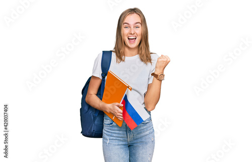 Beautiful young blonde woman exchange student holding russian flag screaming proud, celebrating victory and success very excited with raised arms