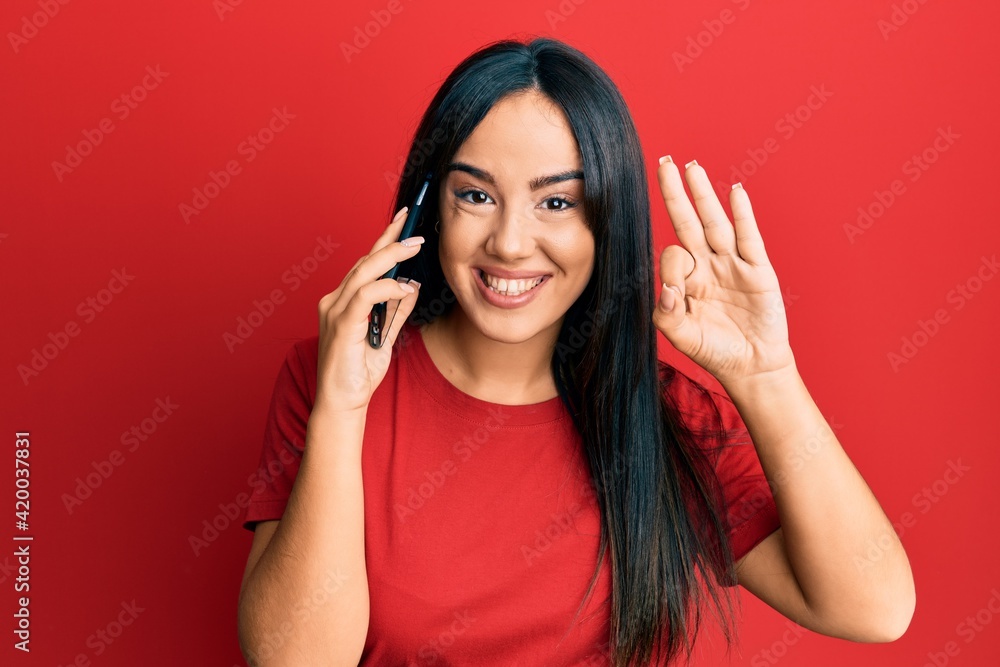 Young beautiful hispanic girl having conversation talking on the smartphone doing ok sign with fingers, smiling friendly gesturing excellent symbol Young beautiful hispanic girl having conversation talking on the smartphone doing ok sign with fingers, smiling friendly gesturing excellent symbol