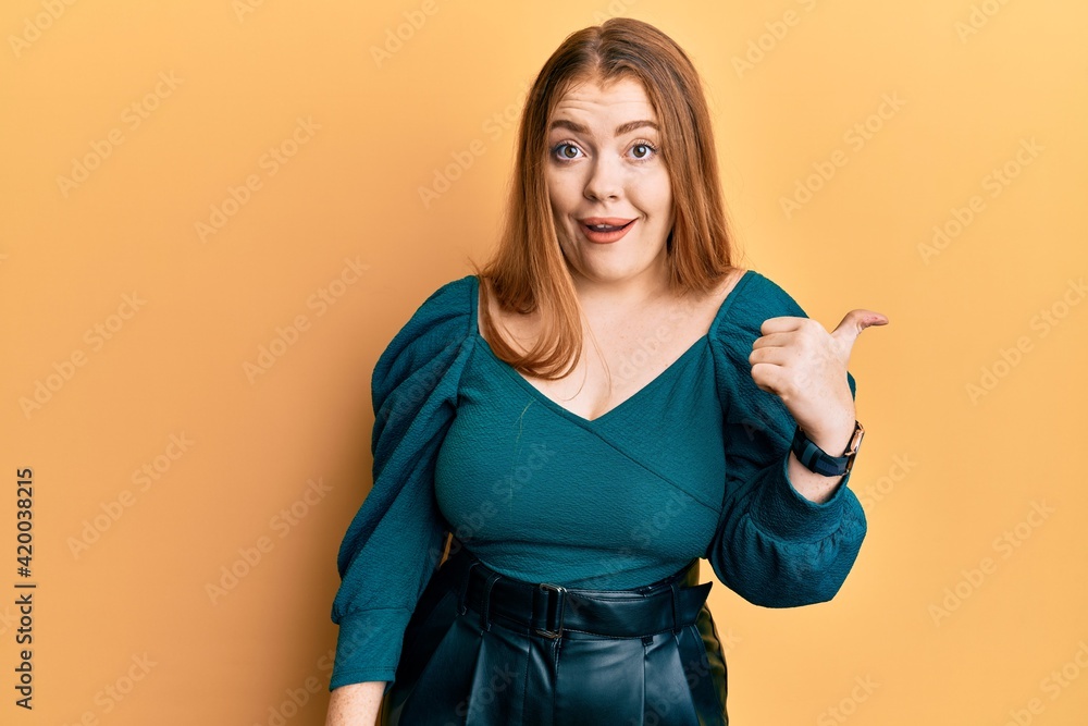 Young beautiful redhead woman wearing elegant and sexy look smiling with happy face looking and pointing to the side with thumb up. Young beautiful redhead woman wearing elegant and sexy look smiling with happy face looking and pointing to the side with thumb up.