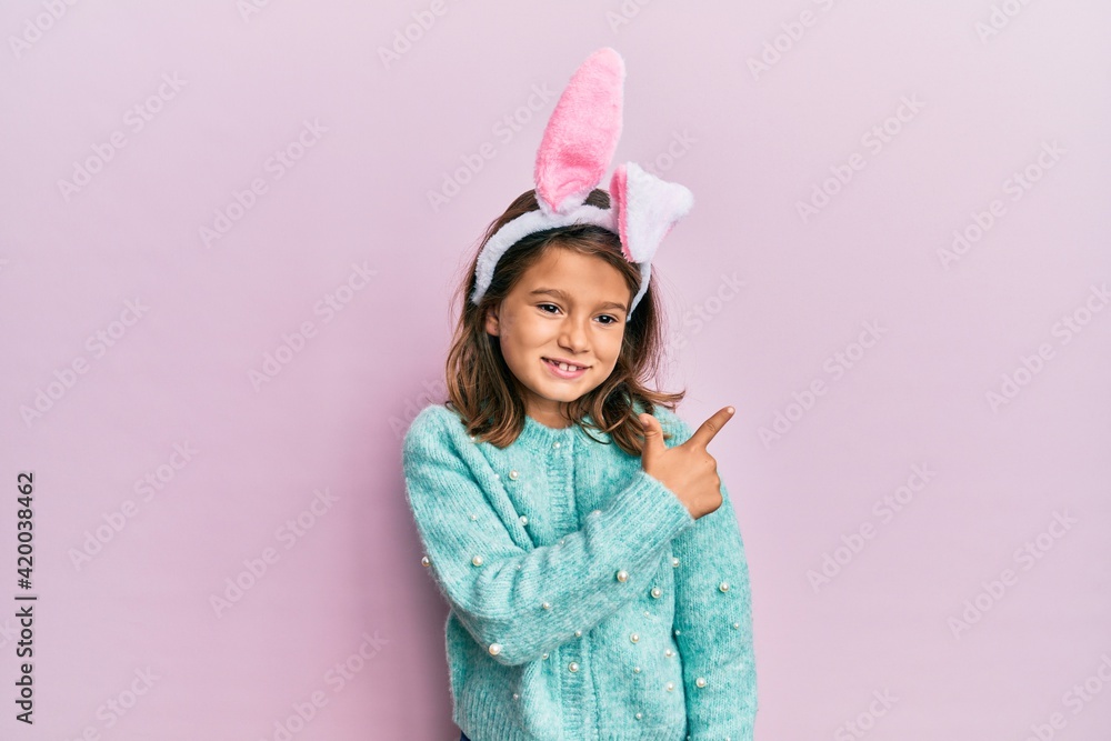 Little beautiful girl wearing cute easter bunny ears cheerful with a smile of face pointing with hand and finger up to the side with happy and natural expression on face