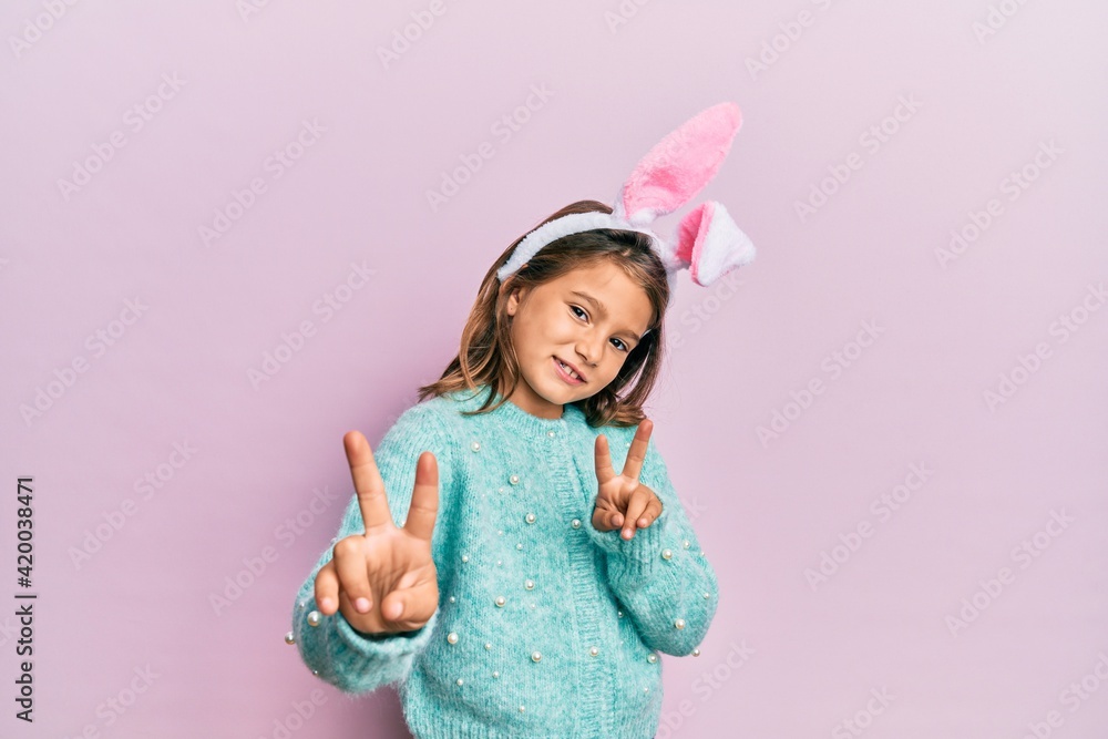 Little beautiful girl wearing cute easter bunny ears smiling looking to the camera showing fingers doing victory sign. number two. Little beautiful girl wearing cute easter bunny ears smiling looking to the camera showing fingers doing victory sign. number two.
