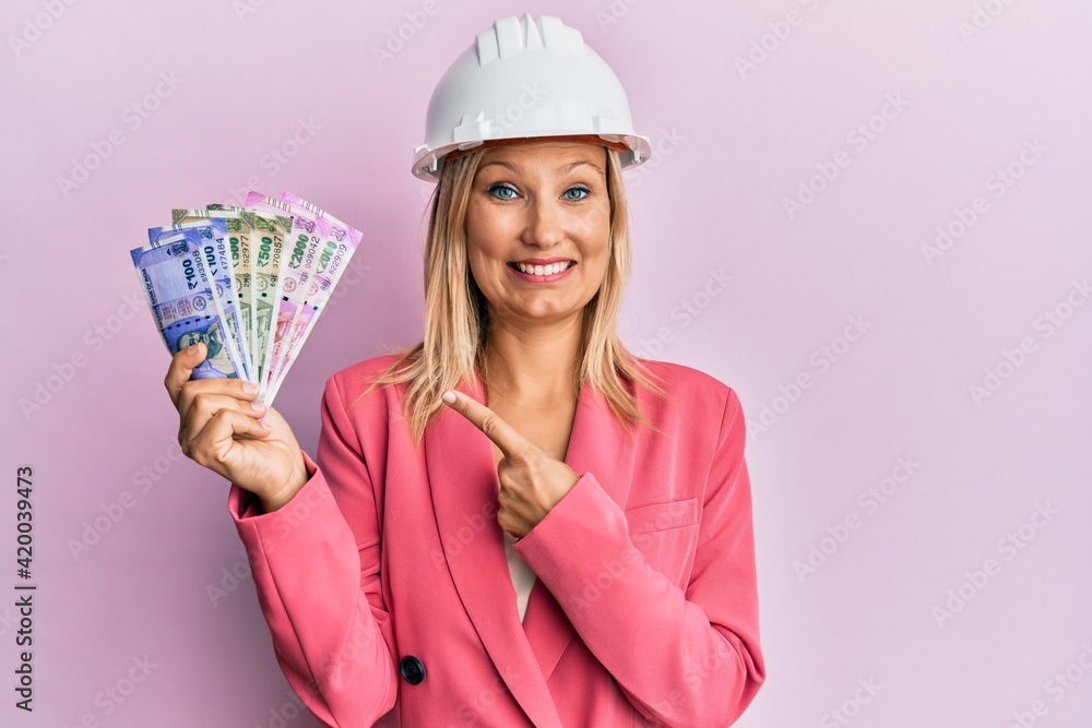 Beautiful middle age blonde woman wearing architect hardhat holding indian rupee smiling happy pointing with hand and finger Beautiful middle age blonde woman wearing architect hardhat holding indian rupee smiling happy pointing with hand and finger