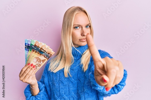 Young blonde girl holding canadian dollars pointing with finger up and angry expression, showing no gesture
