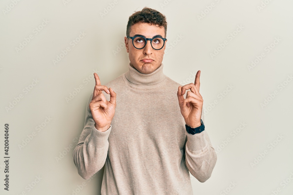 Hispanic young man wearing casual turtleneck sweater pointing up looking sad and upset, indicating direction with fingers, unhappy and depressed. Hispanic young man wearing casual turtleneck sweater pointing up looking sad and upset, indicating direction with fingers, unhappy and depressed.