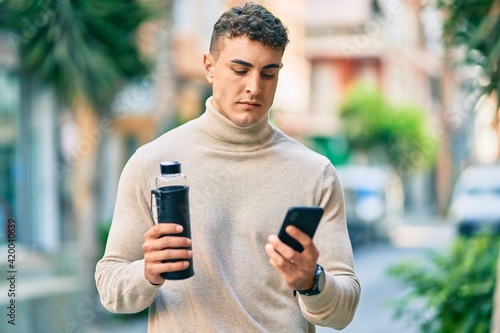 Young hispanic man with serious expression using smartphone and holding bottle of water at the city.