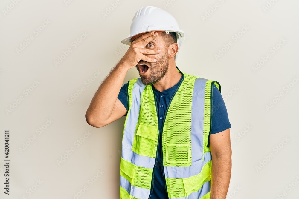 Handsome man with beard wearing safety helmet and reflective jacket peeking in shock covering face and eyes with hand, looking through fingers afraid