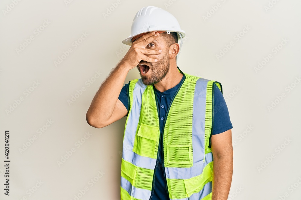 Handsome man with beard wearing safety helmet and reflective jacket peeking in shock covering face and eyes with hand, looking through fingers afraid Handsome man with beard wearing safety helmet and reflective jacket peeking in shock covering face and eyes with hand, looking through fingers afraid
