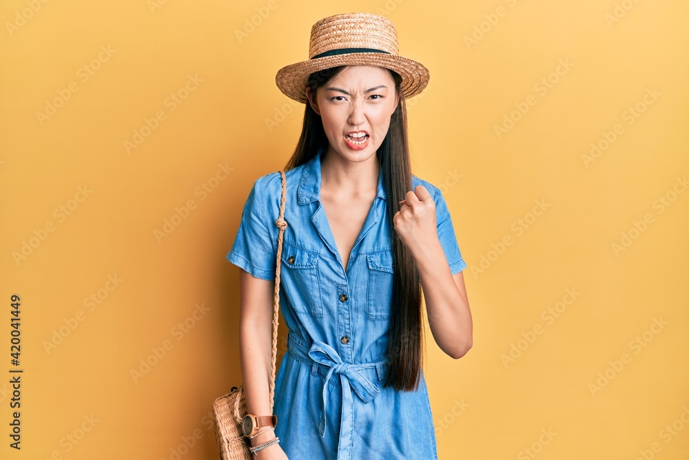 Young chinese woman wearing summer hat angry and mad raising fist frustrated and furious while shouting with anger. rage and aggressive concept.