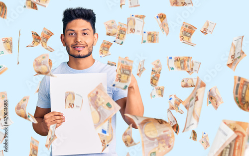 Handsome latin american young man holding cardboard banner with blank space smiling happy and positive, thumb up doing excellent and approval sign