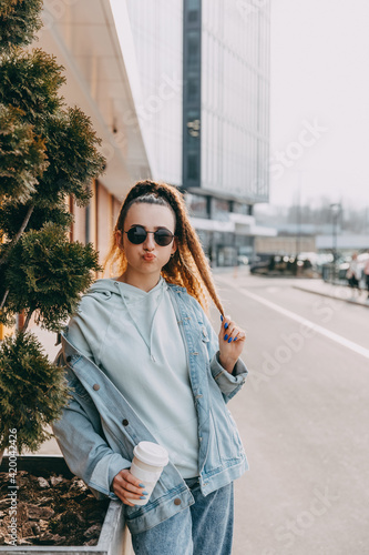 Young woman making funny face, with a reusable cup of coffee, outdoors, on city streets.