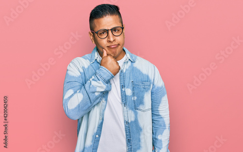 Young latin man wearing casual clothes and glasses serious face thinking about question with hand on chin, thoughtful about confusing idea