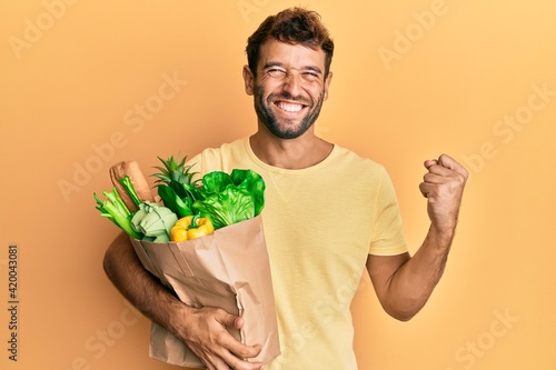 Handsome man with beard holding paper bag with bread and groceries screaming proud, celebrating victory and success very excited with raised arm