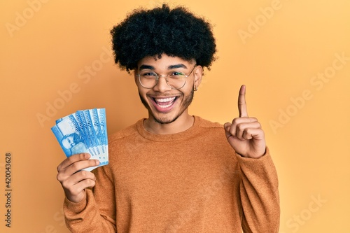 Young african american man with afro hair holding 10000 chilean pesos smiling with an idea or question pointing finger with happy face, number one photo