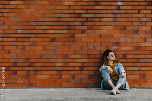 Young woman wearing denim clothes, sitting outdoors on brick wall background.