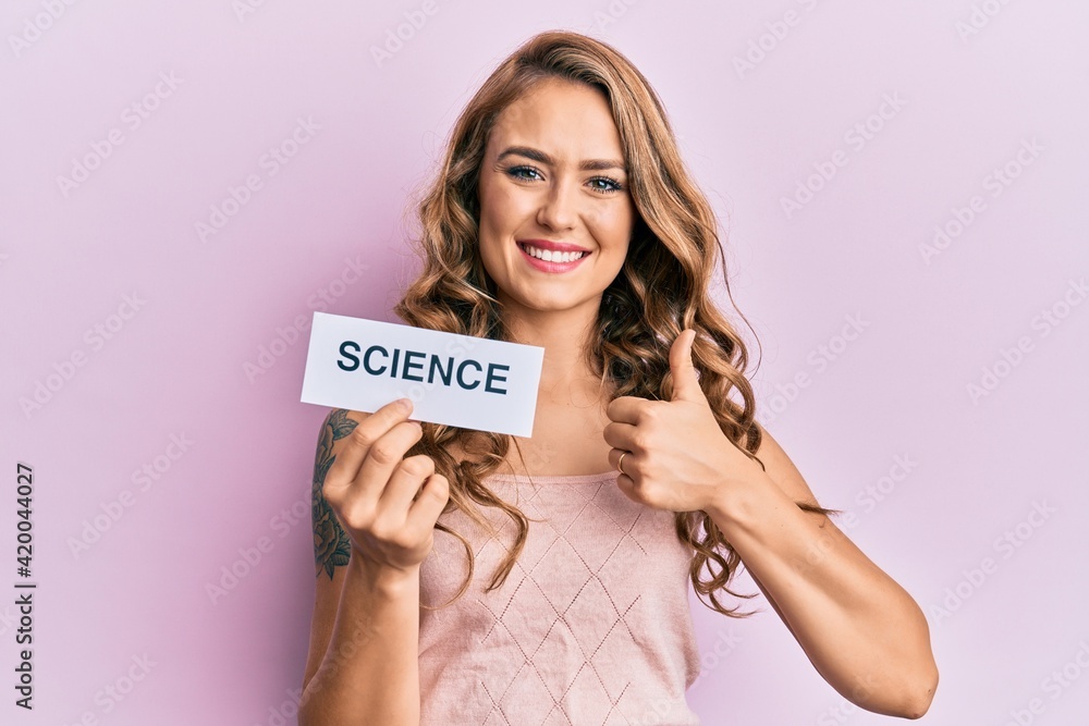 Young blonde girl holding paper with science message smiling happy and positive, thumb up doing excellent and approval sign Young blonde girl holding paper with science message smiling happy and positive, thumb up doing excellent and approval sign