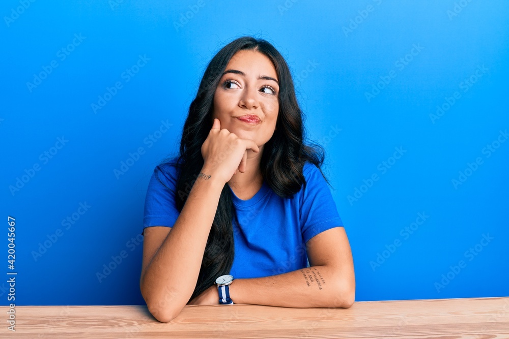 Beautiful brunette young woman wearing casual clothes sitting on the table thinking concentrated about doubt with finger on chin and looking up wondering Beautiful brunette young woman wearing casual clothes sitting on the table thinking concentrated about doubt with finger on chin and looking up wondering