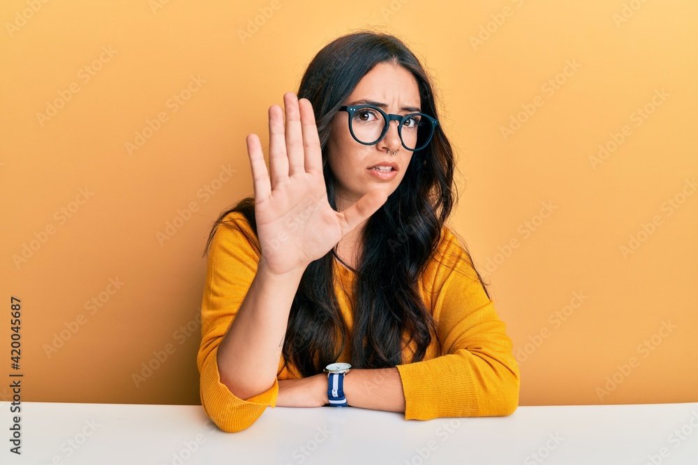 Beautiful brunette young woman wearing glasses and casual clothes sitting on the table doing stop sing with palm of the hand. warning expression with negative and serious gesture on the face. Beautiful brunette young woman wearing glasses and casual clothes sitting on the table doing stop sing with palm of the hand. warning expression with negative and serious gesture on the face.